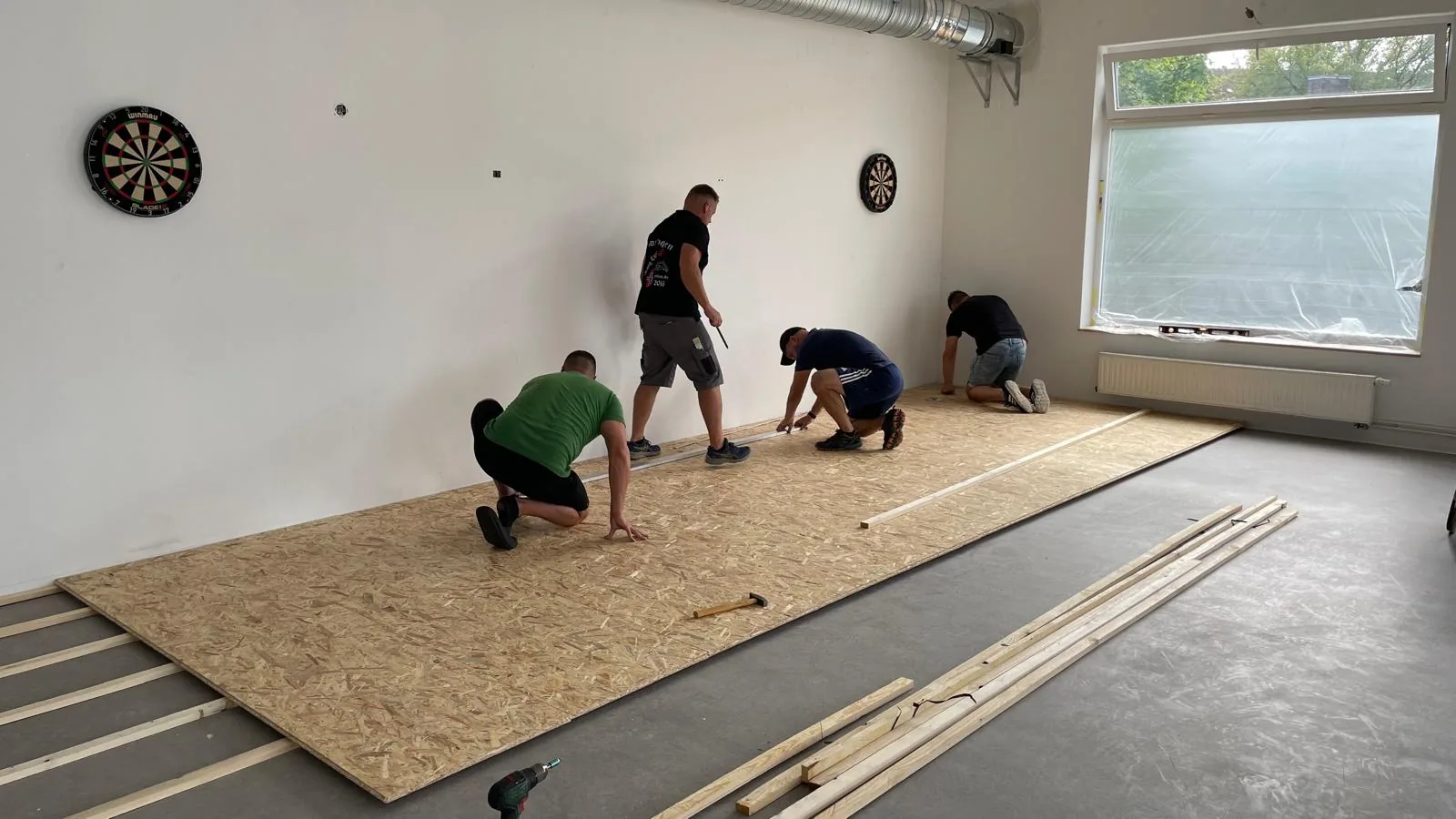 Vier Männer verlegen zusammen eine Holzbodenplatte auf einem Betonboden in einem Raum mit zwei Dartscheiben an der Wand und einem großen Fenster.