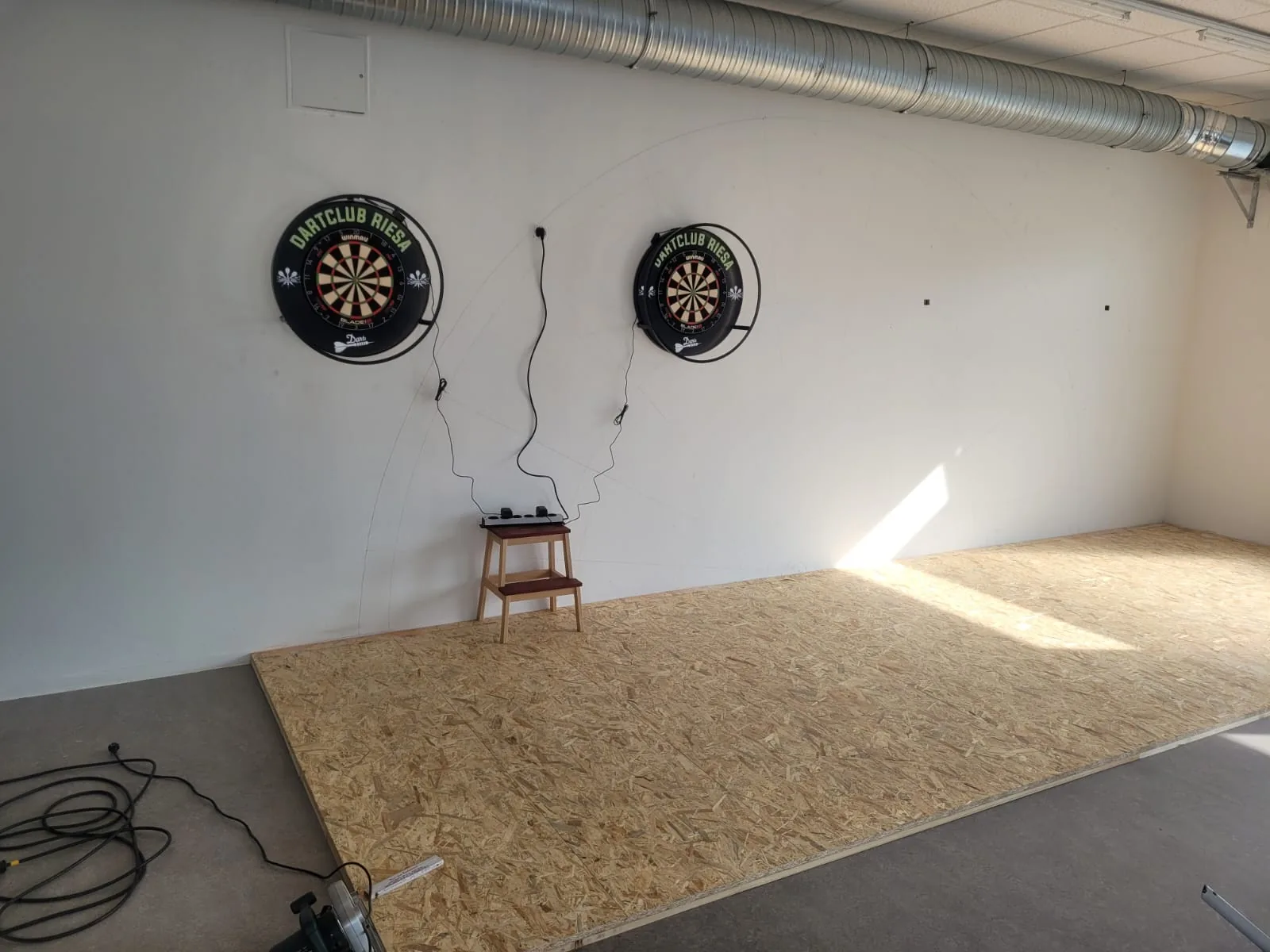 Dartboards mit der Aufschrift Dartclub Riesa an einer weißen Wand, daneben ein kleiner Doppelstufenhocker mit angeschlossenen Kabeln in einem Raum mit OSB-Holzplattenboden.
